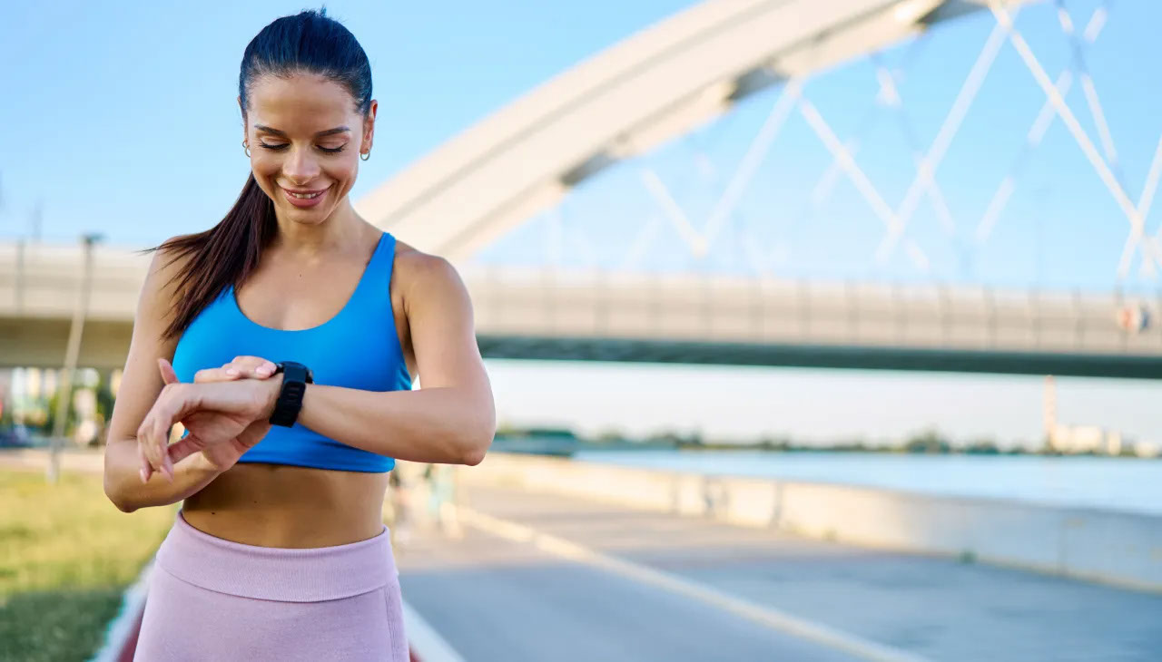 Smiling sportswoman checking her smartwatch while enjoying an outdoor workout near a bridge, embracing a healthy and active lifestyle under the sunny summer sky