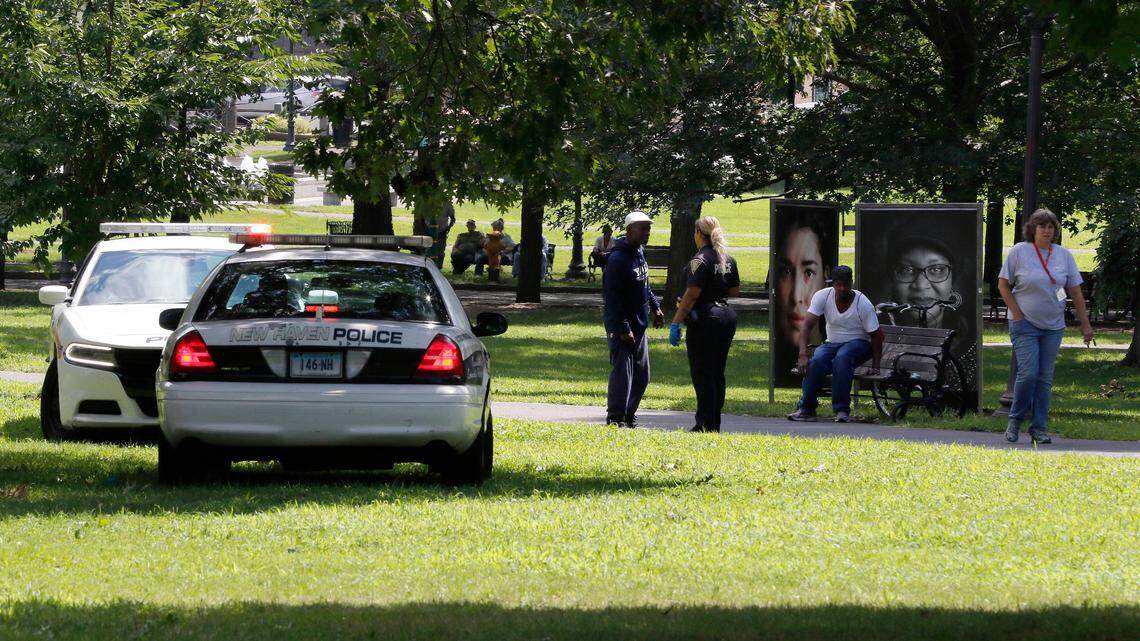 A police officer speaks to a man walking on New Haven Green, Wednesday, Aug. 15, 2018, in New Haven, Conn. A city official said more than a dozen people fell ill from suspected drug overdoses on the green and were taken to local hospitals. (AP Photo/Bill Sikes)