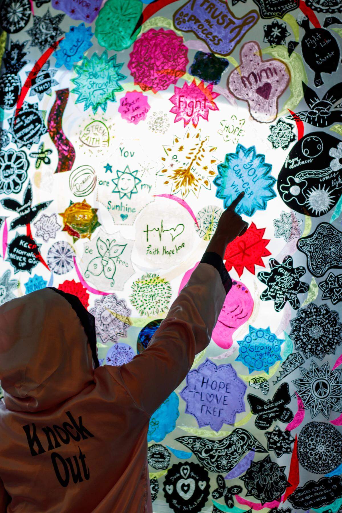 El Instituto Oncológico de Miami tiene una ventana cubierta de obras de arte creadas por pacientes de cáncer. Latoya Simmons, de 42 años, sobreviviente de cáncer de mama, mientras lleva la bata rosa que le sirvió de armadura durante la quimioterapia, señala una de las obras que hizo mientras recibía tratamiento en el hospital.