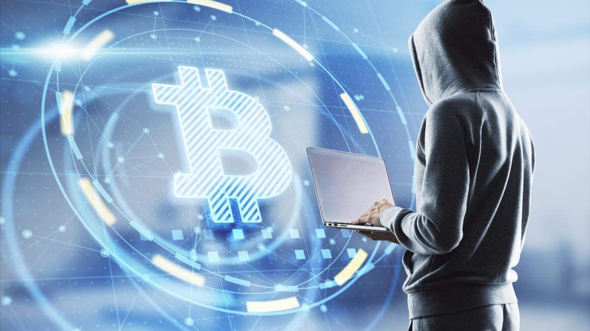 Hacker in hoodie using laptop computer with glowing Bitcoin interface in blurry office interior.