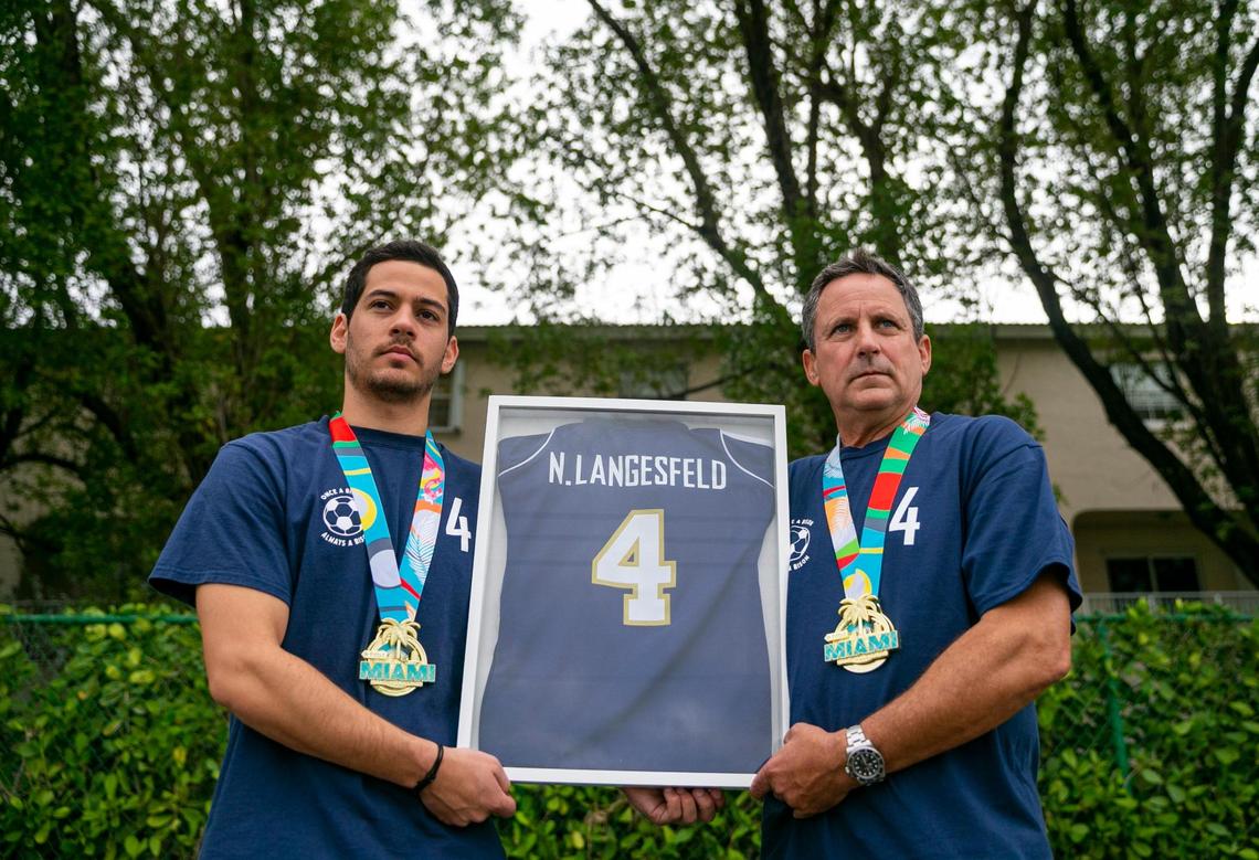 Martin Langesfeld, 24, and his father, Pablo Langesfeld, 56, hold a framed high school soccer jersey belonging to Nicole Langesfeld at the Turnpike Trail in Doral, Florida, on Wednesday, January 26, 2022. Both Martin and Pablo were planning to run in the Miami Marathon in honor of Nicole and her husband, Luis Sadovnic, both of whom were killed in the Surfside condominium collapse.