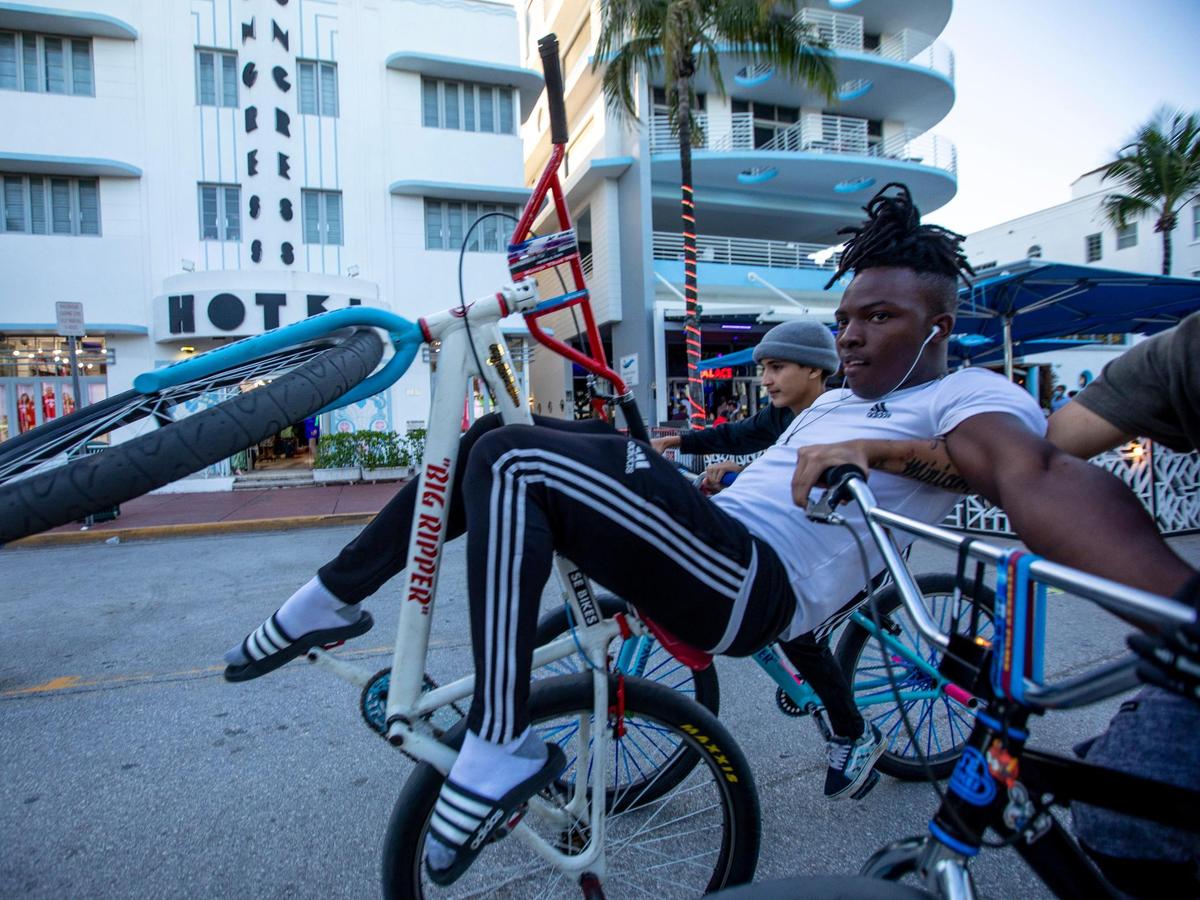 A teenager props himself up while he pops a wheelie and balances on his friends’ bikes as they coast south down Ocean Drive before curfew in Miami Beach, Florida, on Tuesday, March 23, 2021.