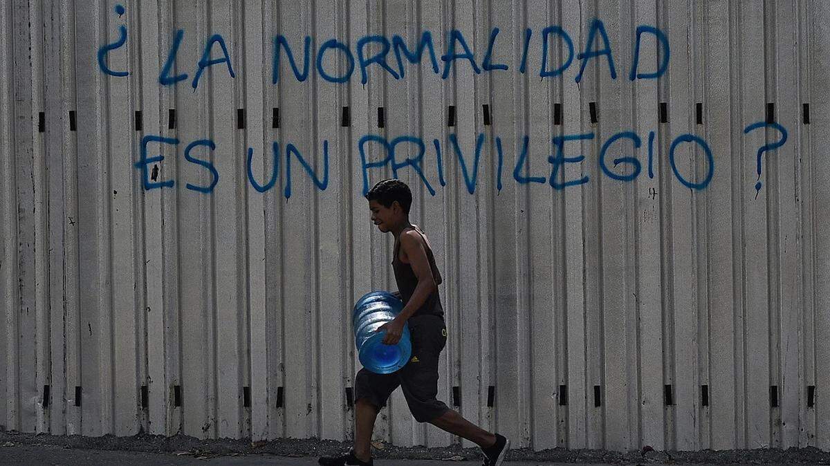 A boy carries a drum of water as he walks past graffiti reading “Is Normality a Privilege?” during a power outage in Caracas ini 2019.