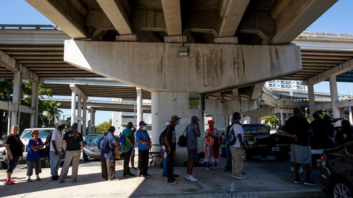 Houseless people wait in line under the highway as volunteers help pass out donated clothing and food during a donation and feeding event hosted by The Smile Trust, Inc. near Southwest 2nd Avenue and Second Street in Downtown Miami, Florida, on Sunday, October 17, 2021. The Smile Trust, Inc. is an International Nonprofit Organization that fights against homelessness and works toward fixing food insecurities.