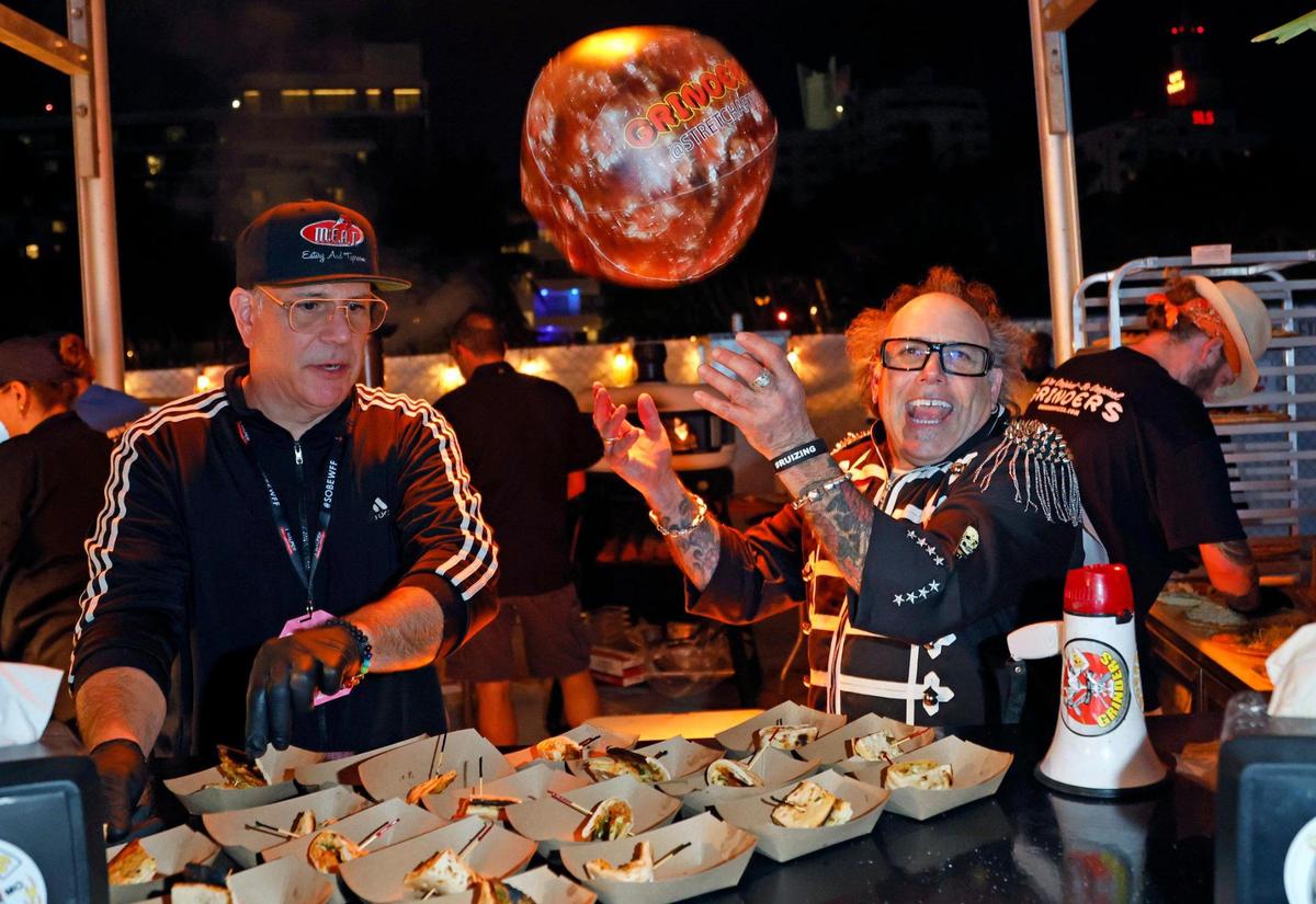 George Patti, left, serves up pizza as Stretch Grinder tosses a Grinders beach ball during the South Beach Wine & Food Festival on the sand by the Ritz Carlton in Miami Beach, Florida on Saturday, February 25, 2023.