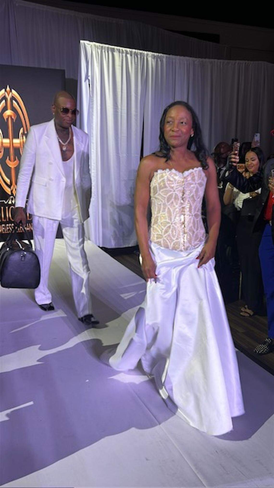 Haitian designer Phelicia Dell, dressed in one of her designs, walks the runway of her fashion show on Saturday, April 4, 2026 at Gulfstream Park. Behind her is with Jean Herard “Richie” Richard, lead drummer of the Haitian band Klass, with whom she designed a duffel bag (in his hands).