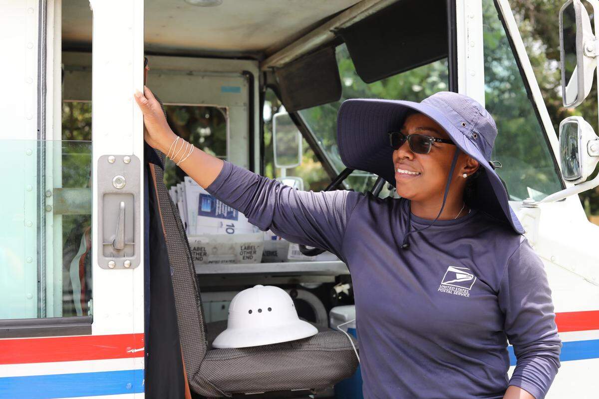 Olivia Gordon, a mail carrier for the United States Postal Service, stands for a portrait alongside her un-air-conditioned mail truck, wearing the floppy hat and long-sleeved shirt that keeps her out of the sun, on July 5, 2023.