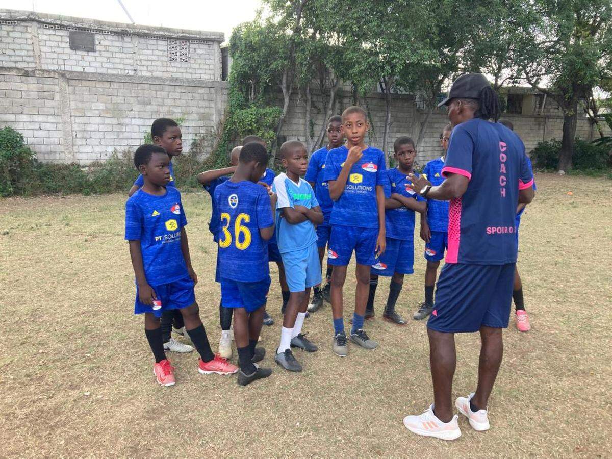 Eleven-year-old Josué St. Vilus, in blue, was a goalkeeper for Olympique École de Football in Sarthe, Haiti who dreamed of one day defending the country’s red-and-blue national colors. He was killed on Saturday, May 16, 2026 when a stray bullet tore through his abdomen as his family tried to flee gang attacks.