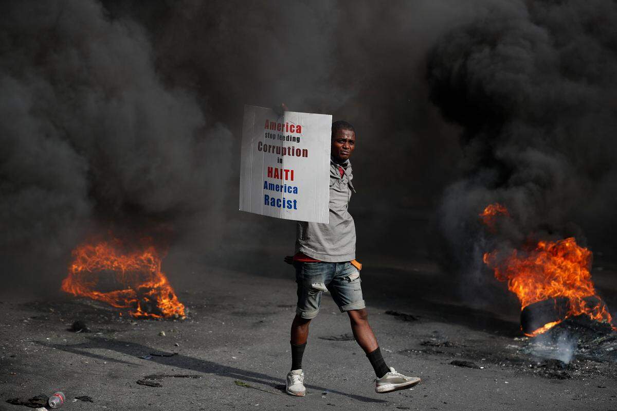 A protester carries a sign directed at the U.S. as he walks between burning tires in Port-au-Prince, Haiti, Friday, Oct. 4, 2019.