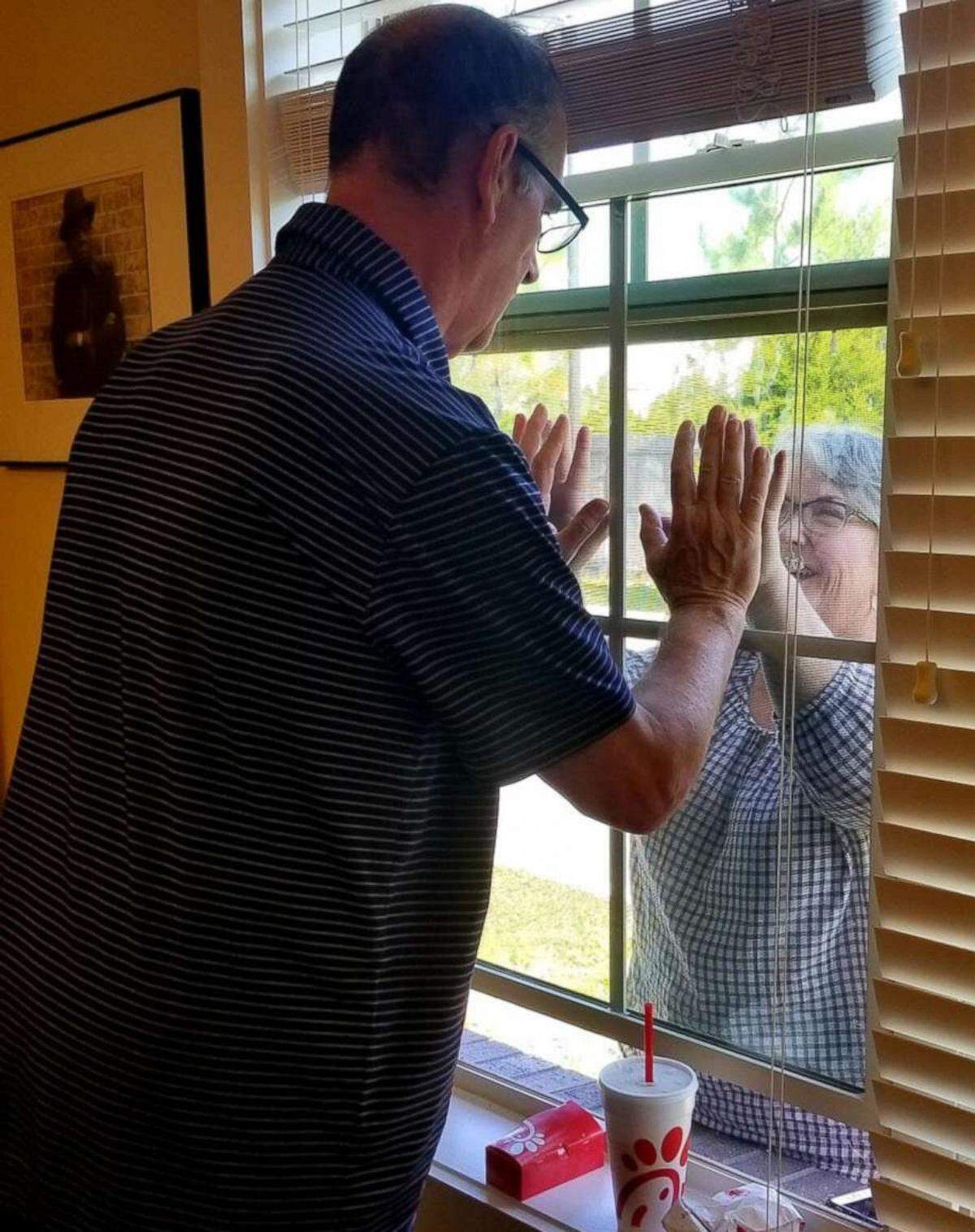 Mary Daniel and her husband Steve join hands through a window at the Rosecastle Assisted Living and Memory Care facility in Jacksonville. Desperate to see her husband in person, Daniel took a part-time job as dishwasher and started a national Facebook group, Caregivers for Compromise, to advocate for changes to restrictive visitation policies.