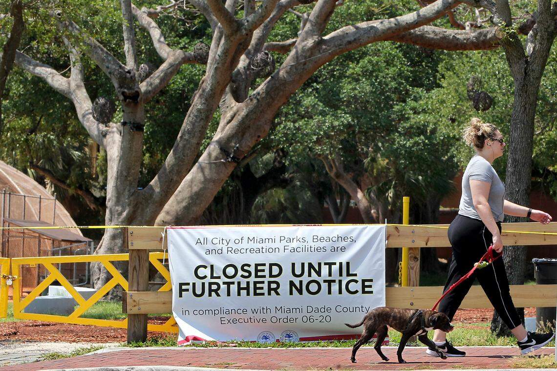 A woman walks her dog on the sidewalk as all city of Miami parks remained closed, including the Peacock Park in Coconut Grove, until further notice due to the COVID-19 pandemic, on Tuesday, April 14, 2020.