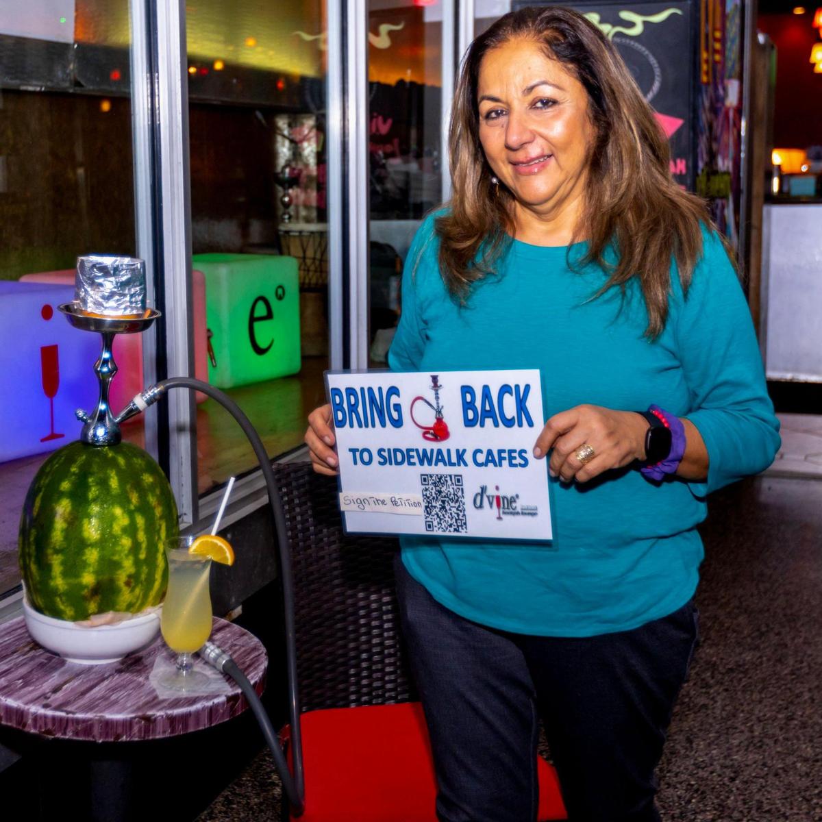 Claudia Herman, owner of D’Vine Hookah Lounge, holds up a sign that reads “Bring Hookah Back to Sidewalk Cafes” in front of her business on Lincoln Road in Miami Beach on Dec. 1, 2022. Herman is fighting a city ban on serving hookah at sidewalk cafes on public property.