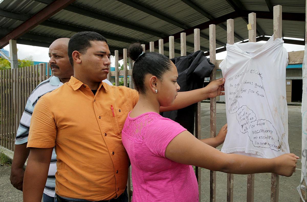 Student Genesis Velázquez, with her father, Felipe Velázquez (far left), and her cousin Jafenier Velázquez, reads pleas from students to save their school written on uniforms. The messages were meant for Puerto Rico’s Education Secretary Julia Keleher, asking her to reopen Marcos Sánchez, a school recently closed after Hurricane Maria.  The school, said Genesis’ father, “was the only thing here to unite the community. The only thing.”