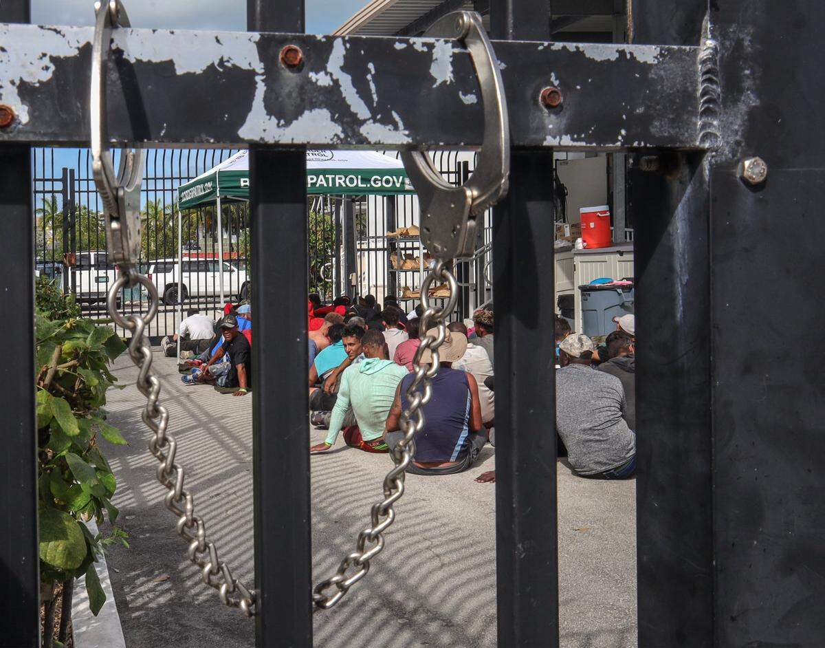 A pair of handcuffs are secured on the exterior gate as Cuban migrants sit on the ground under the midday sun on Wednesday, Jan. 4, 2022, while waiting to be processed at the Border Patrol station in Marathon in the Florida Keys. They are among hundreds of people from Cuba, and one large group of over 100 men and women from Haiti, who’ve landed across the island chain since the end of last week.