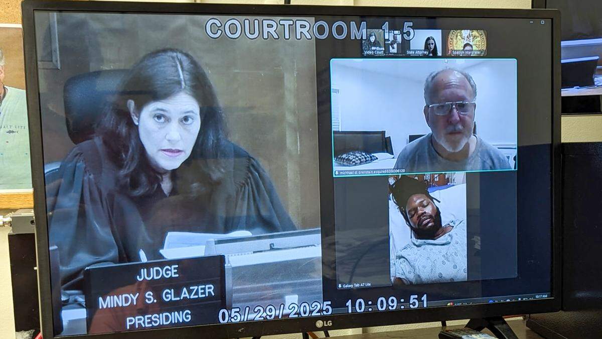 Miami-Dade Circuit Court Judge Mindy Glazer holds a bond hearing Thursday morning, May 29, 2025, over Zoom in the Jackson Memorial Hospital room of Menelek Emmanuel Clarke. Clarke was the driver who was shot by a Miami policeman after hitting him in his car outside a Caribbean music festival at Bayfront Park on Sunday night. Clarke is in the hospital bed; his lawyer his attorney Michael Orenstein is pictured above Clarke, who was charged with four felonies