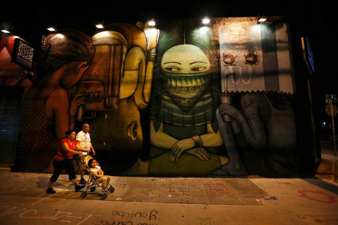 A couple pushing a child in the stroller walk past a masked mural painting on the Wynwood Kitchen & Bar restaurant. The normally bustling Wynwood arts district was void of its usual crowds Wednesday evening, April 1, 2020, as Miami-Dade residents were following orders to stay home.