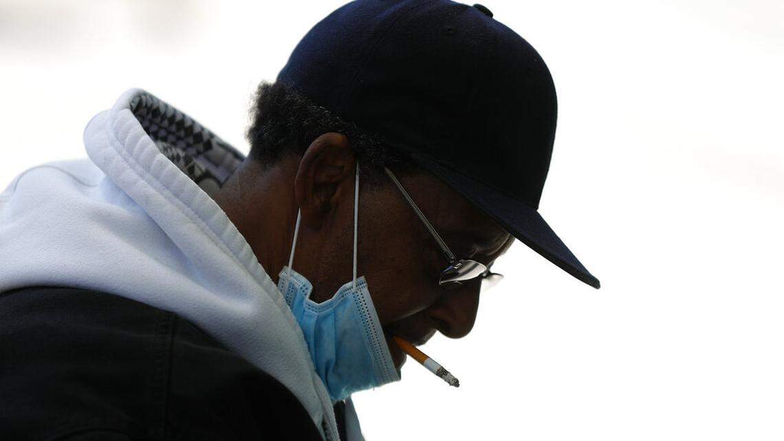 A man smokes a cigarette while wearing a protective mask while waiting for a bus in Detroit, Wednesday, April 8, 2020. Detroit buses will have surgical masks available to riders starting Wednesday, a new precaution the city is taking from the new coronavirus COVID-19. (AP Photo/Paul Sancya)