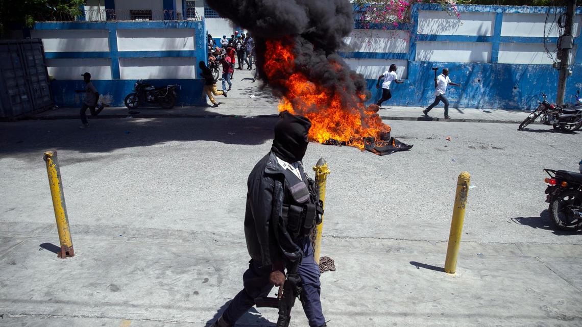 A masked police officer walks with his weapon past a barricade set alight in front of a police station in Delmas, during a protest by a disgruntled sector of the Haitian police force known as Fantom 509, in Port-au-Prince, Haiti, Wednesday, March 17, 2021. The protests started when officers and police academy cadets marched toward police headquarters to demand that the bodies of several officers killed during a raid last week on the Village of God shantytown be recovered from the gang still holding them.