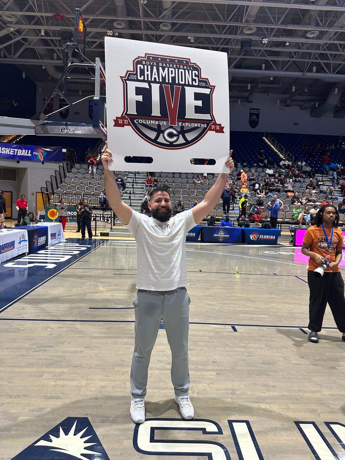 Columbus boys’ basketball coach Jorge Milo holds up a sign commemorating the Explorers’ fifth consecutive state championship, which tied a state record. The Explorers beat Sarasota in the 7A final on Saturday at UNF Arena in Jacksonville, Fla.