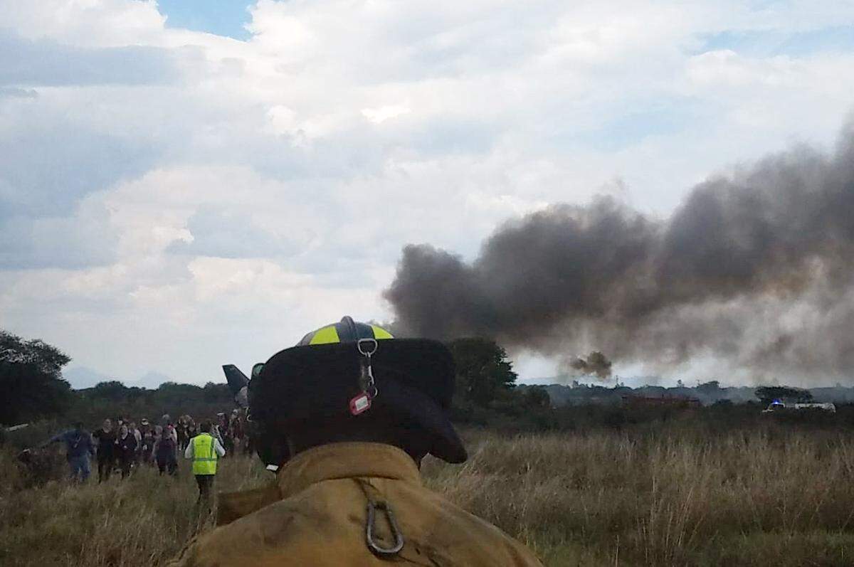 Smoke billows from a downed Aeromexico jetliner as passengers walk away from the crash site in a field near the airport in Durango, Mexico, Tuesday, July 31, 2018. Durango state Gov. Jose Aispuro said all 99 passengers and four crew members made it off the plane, but the pilot was severely injured.