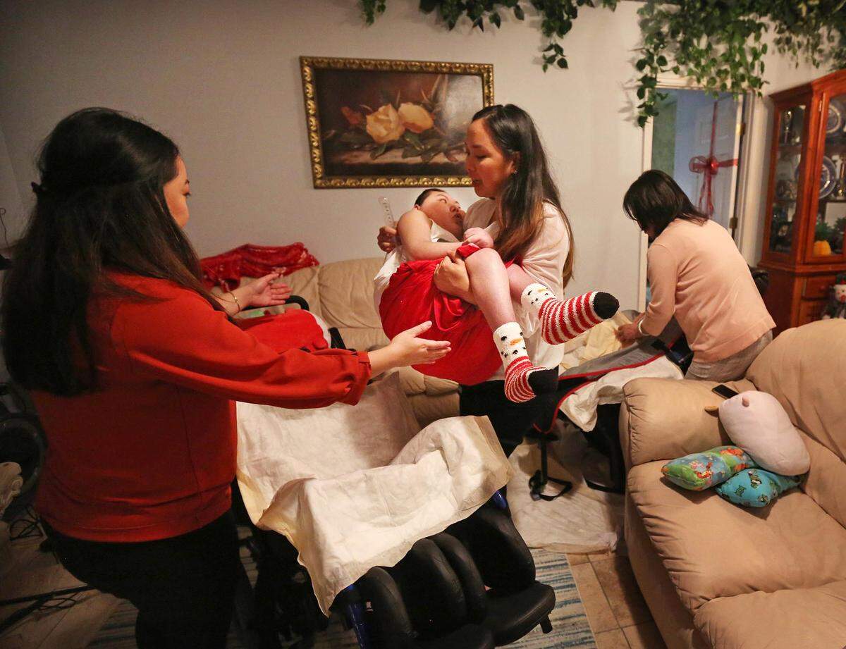 Justin Nguyen, 24, is carried by his sister Jennifer Pham to his wheelchair as their older sister, Jessica Pham, reaches out to assist her. Their mother, Julie Nguyen, works on Justin’s medical equipment in the background.