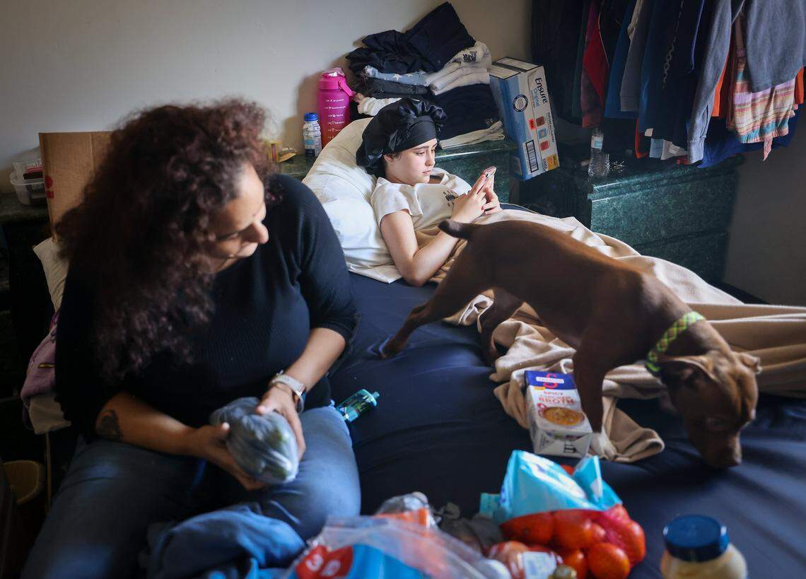 Maylynda, right, scrolls through TiK Tok videos as her pet, “Hazel” roams around the bed as Diana Olivo looks on. Michael Torres, Diana Olivo, Maylynda, and her two brothers have been sharing a motel room for the last six months. He does not know how long they'll be able to afford to stay there, and he reflects on the challenges of being unsheltered on Wednesday, February 25, 2026, in Miami-Dade, Florida.