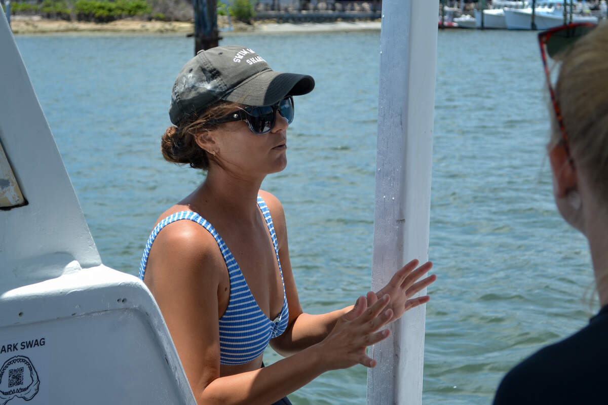 Sporting a gray “swimming with sharks” baseball cap, Jordan Lempke, an employee for Florida Shark Diving in Jupiter, Florida, explains to tourists what will happen once the group sails five miles off the coast to dive with sharks on June 12, 2026.