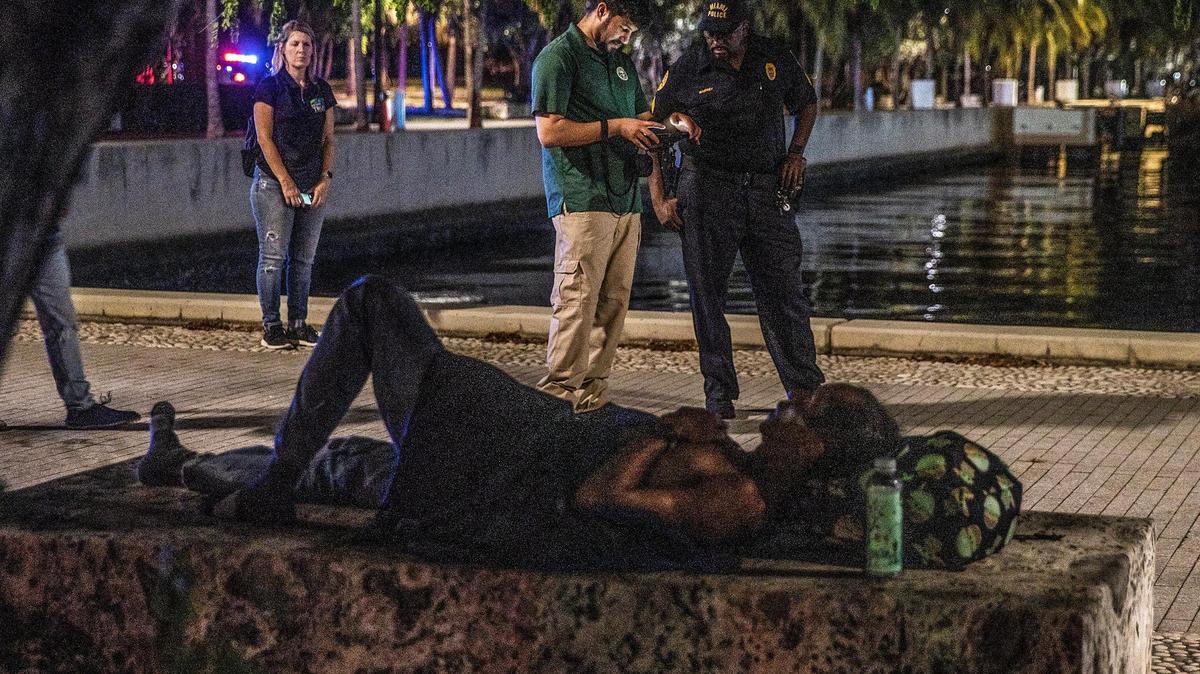 Un desamparado duerme en un banco en el Museum Park, mientras que los especialistas municipales de Miami Christian Candelier (izquierda) y Ricky Leath, caminaban alrededor del parque durante el censo anual de desamparados de verano Point-in-Time (PIT) del Condado Miami-Dade, donde diferentes equipos realizaron el recuento anual de verano de los indigentes desde Homestead y Miami Beach hasta North Miami, el jueves 18 de agosto de 2022.