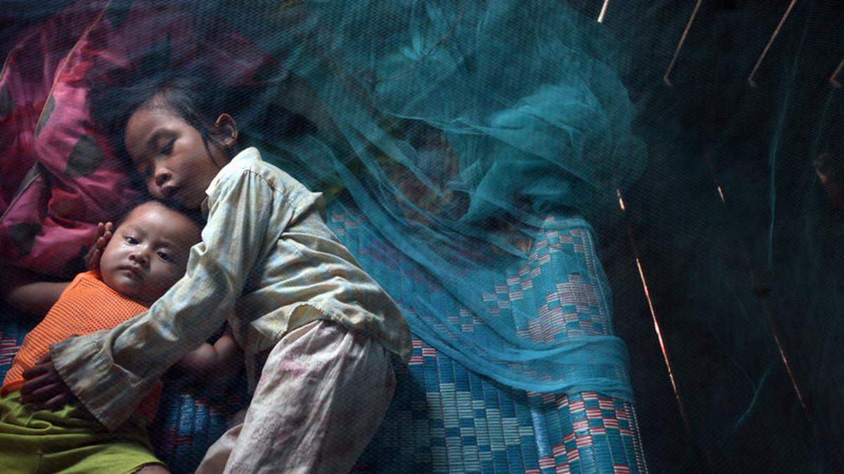 Two Cambodian children lie in a bed with a net, which protects them from mosquitoes carrying malaria.