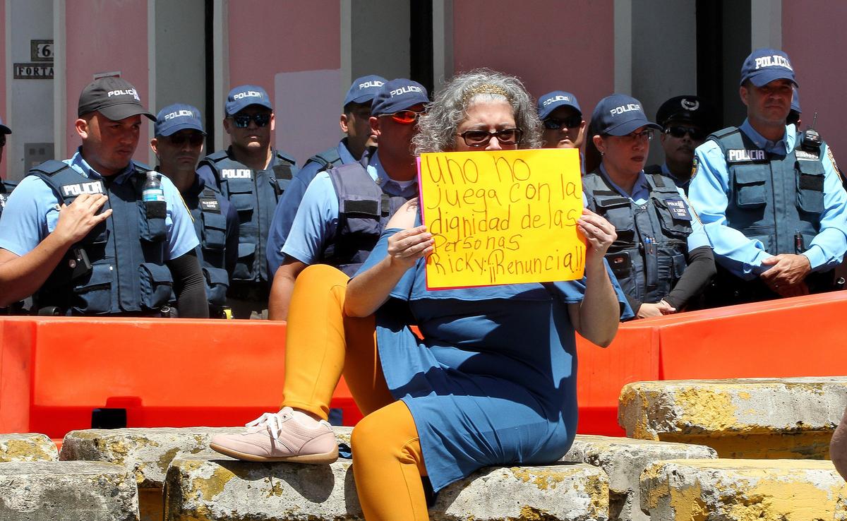 Jeannette Saldana, in front of a security perimeter set up by police in Old San Juan, displays a sign demanding the resignation of Gov. Ricardo “Ricky” Rossello.