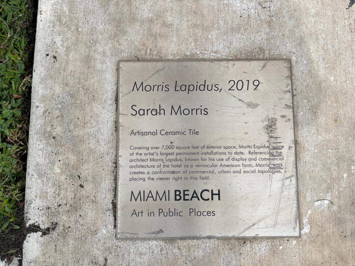 La placa de Morris Lapidus". obra de Sarah Morris, permanece en la acera frente al muro este del Centro de Convenciones de Miami Beach. La obra de arte del muro este estaba en tan mal estado que la ciudad instaló una cubierta protectora opaca sobre ella. Amanda Rosa