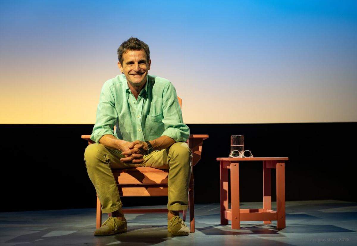 The set is purposefully sparse: an Adirondack chair and table on stage left, six-cylinder lights positioned in the corners, and a large LED screen stretching across the back wall.