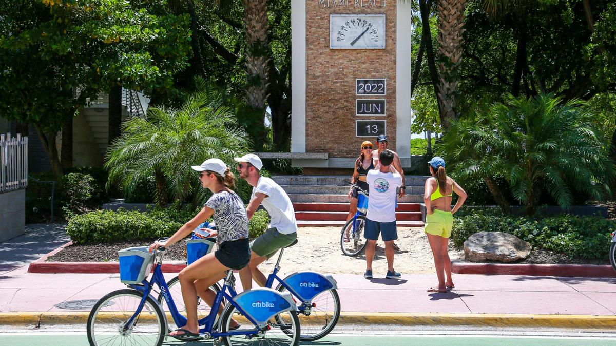 A couple of people ride their bikes while others take a photo in front of The Clock Thermometer on Ocean Drive and 10th Street as temperature rises above 90 degrees Celsius in Miami Beach, Florida, on Monday, June 13, 2022.