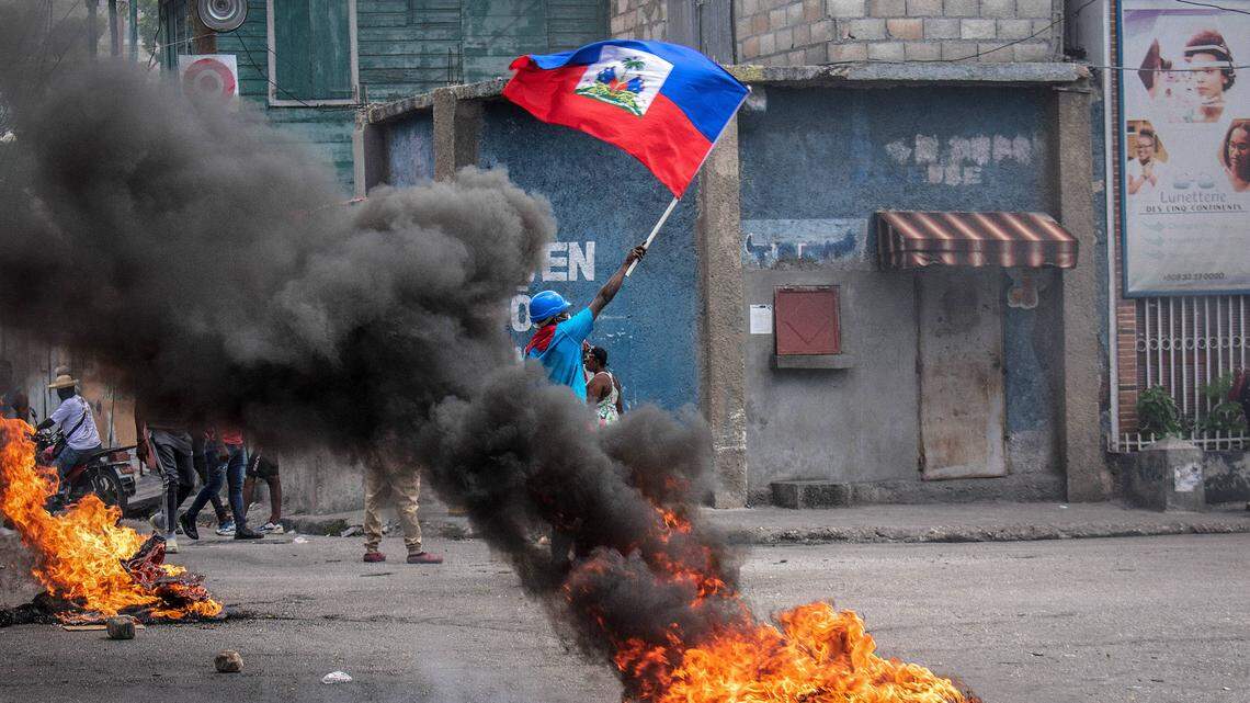 In a photo from Auf. 14, 2023, a man with the Haitian flag in hand stands in the middle of a fire to ask for peace. Haiti has moved closer to total collapse in recent days.