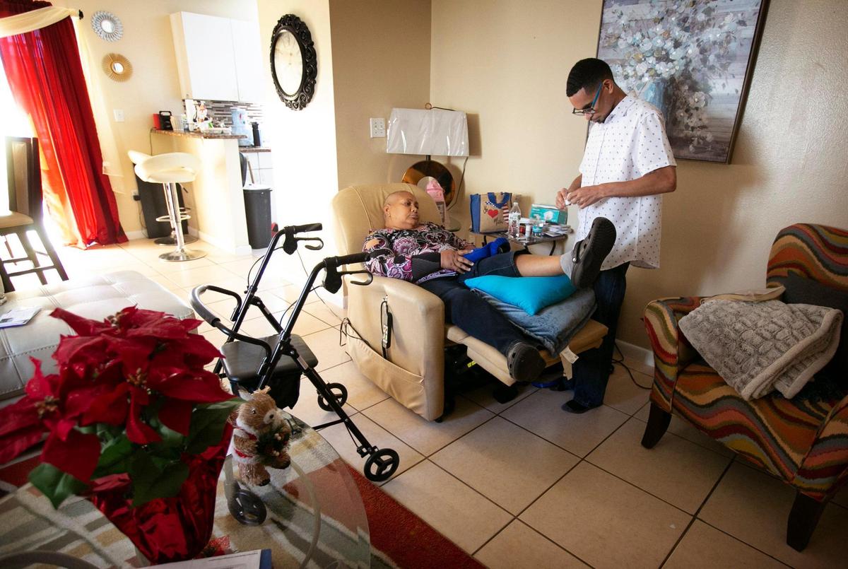Andre Amador helps his mother, Maria Rosario, get settled into a chair that helps her to rest. Rosario was diagnosed with breast cancer in 2015 and the cancer has now spread to her back and her bones.