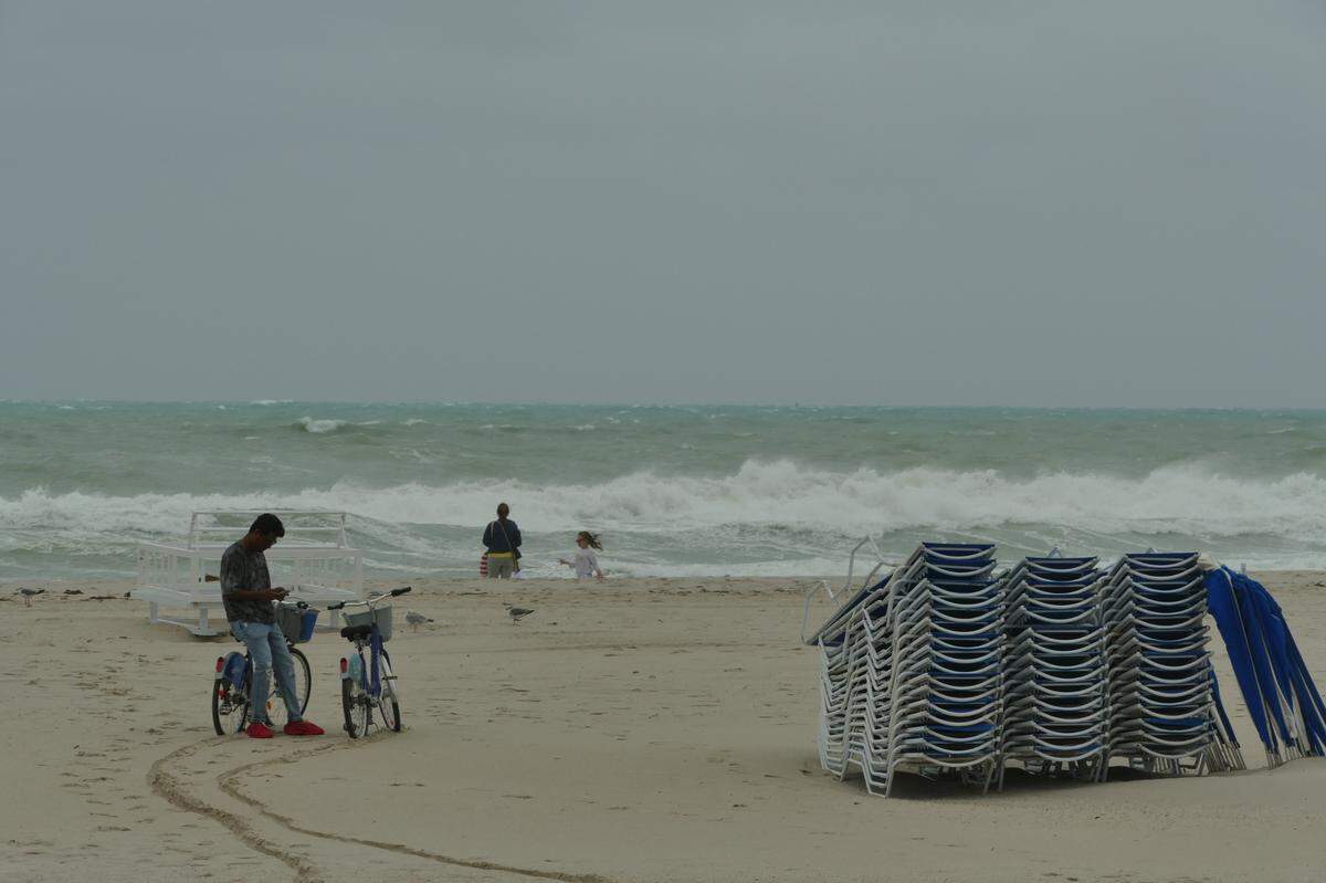 A man waits next to two rented bikes as a woman off-camera takes a closer look at the rowdy waves in Miami Beach, Florida, on Friday, Dec. 15, 2023.