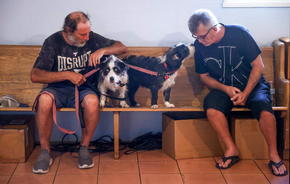Bill Ledger, 54, left, sits with his dogs - “Scappy,” and “Scavenger” while waiting for lunch to be served as volunteer Freddy Clements, 61, interacts with the beloved pets on Monday, January, 26, 2026, in Oakland Park, Florida. Father Bob Caudill at All Saints Catholic Mission in Oakland Park fed the homeless every day, despite city officials claiming it was against the rules. The city fined him $150 each day since 2016. He sued the city for religious discrimination. The trial was set to begin soon.