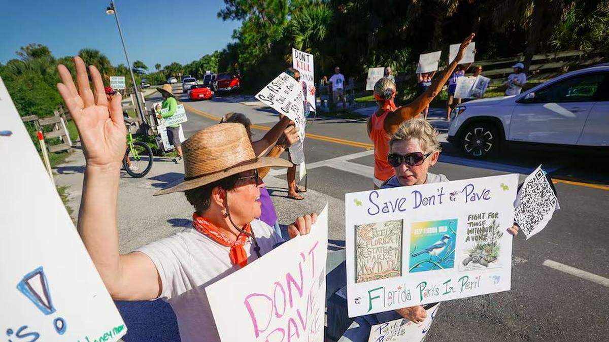 Michelle Birnbaum, 59, left, and Kathie Nutter, 67, right, both of Dunedin, hold signs and wave to drivers at the entrance to Honeymoon Island State Park during a rally Sunday, Aug. 25, 2024 in Dunedin.
