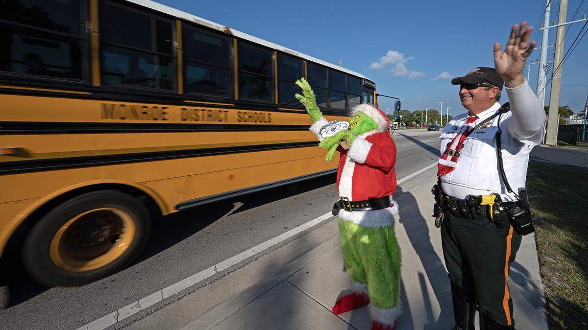 Monroe County Sheriff’s Office Colonel Lou Caputo, playing the Grinch, and Deputy Andrew Leird, wave at a school bus rolling on the Florida Keys Overseas Highway Tuesday, Dec. 13, 2022, in Marathon.