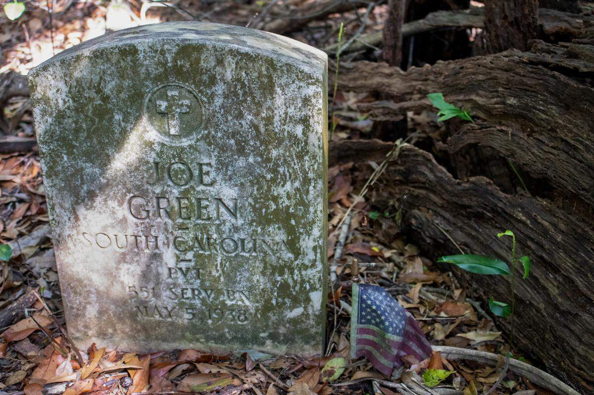 The headstone of Joe Green, a military veteran, sits adjacent to a faded American flag and a dead tree lying on its side in the Oak Tree Union Colored Cemetery of Taylorville in Groveland, Fla., on Friday, March 25, 2022. The cemetery that dates to 1895 has become overgrown with vegetation but still has multiple headstones visible.