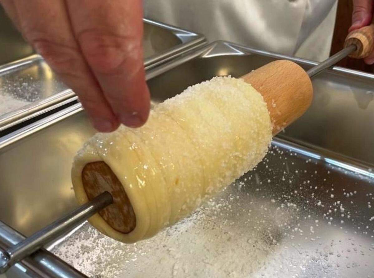 A baker sprinkles sugar on the dough of a chimney cake.