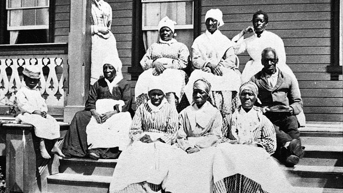 A group of women and a man, presumably enslaved, sit on the steps of the Florida Club, St. Augustine, Florida, mid 19th Century. A white woman, possibly a manager or overseer, stands behind them to the left and a child sits on the newel post. (Photo by Hulton Archive/Getty Images)