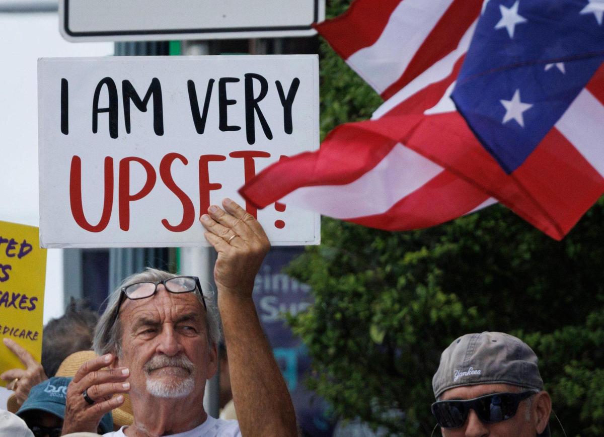 A man holds a sign and an upside down American flag flies in front during a May Day rally themed “Protect Social Security, Protect Workers” on Thursday, May 1, 2025, outside of the Social Security Administration building in Miami.