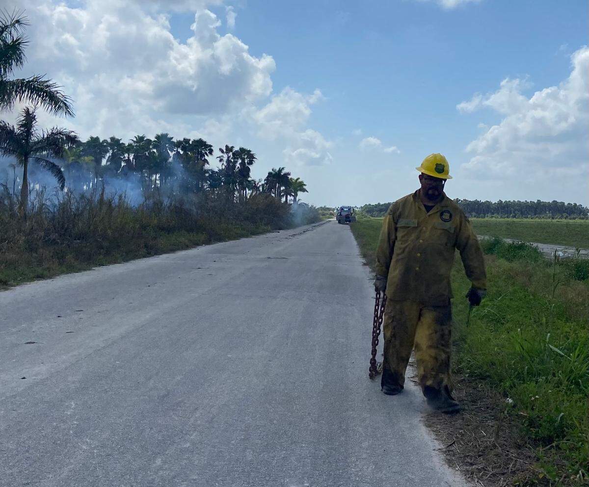 Florida Forest Service Firefighter Luis Pineda carries a chain along the roadside as a fire smolders in the background at Southwest 112th Avenue and 268th Street Friday, March 3, 2023.