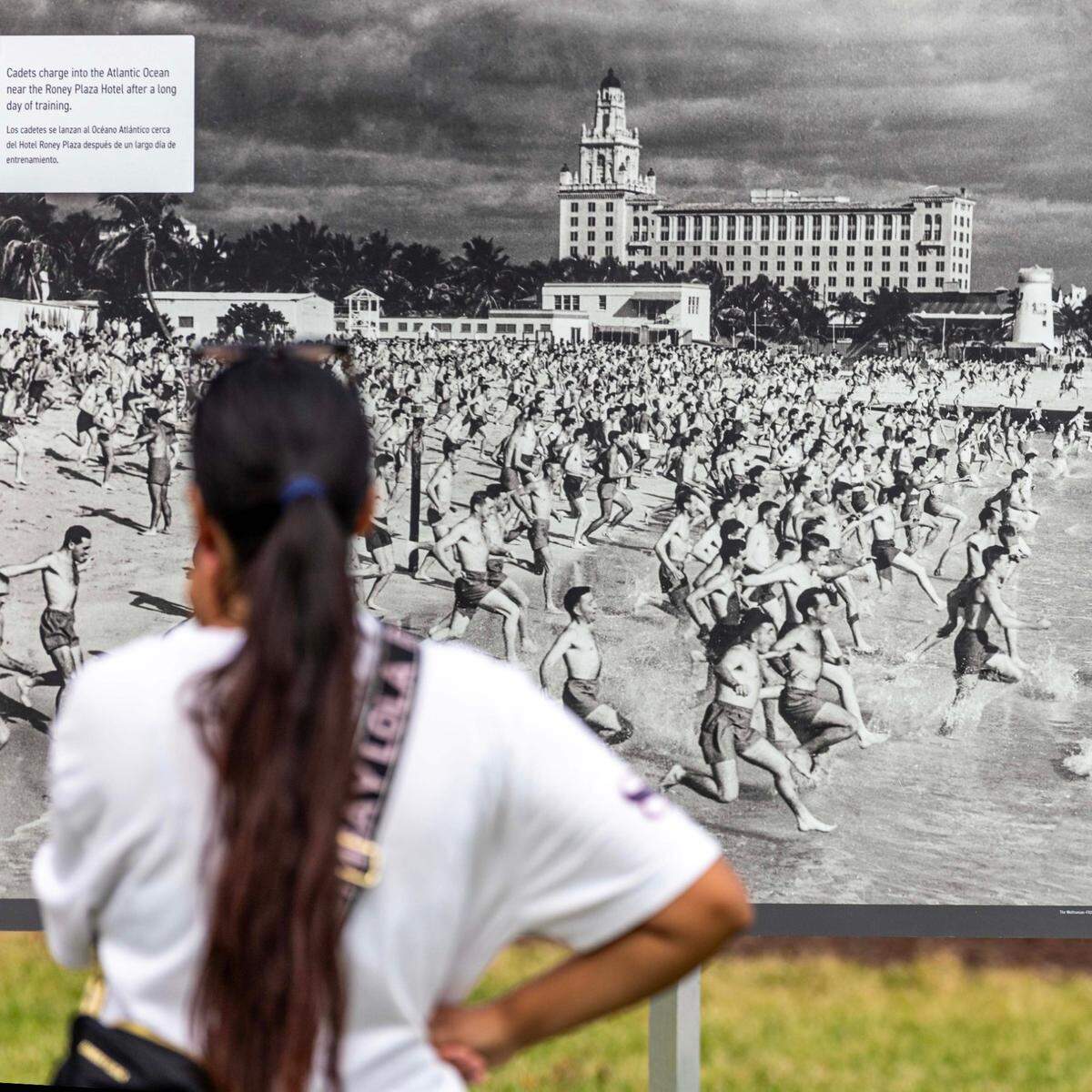 A woman looks at a photo showing cadets take a swim after a day of training from the temporary outdoor exhibition: “Boot Camp in Paradise: World War II in Miami Beach,” featuring over 150 photographs and printed materials from 1942-1945, when Miami Beach hosted over 600,000 U.S. troops training for combat, during the 16th Annual Veterans Day Parade off Ocean Drive on Monday, Nov. 11, 2024, in Miami Beach, Fla.