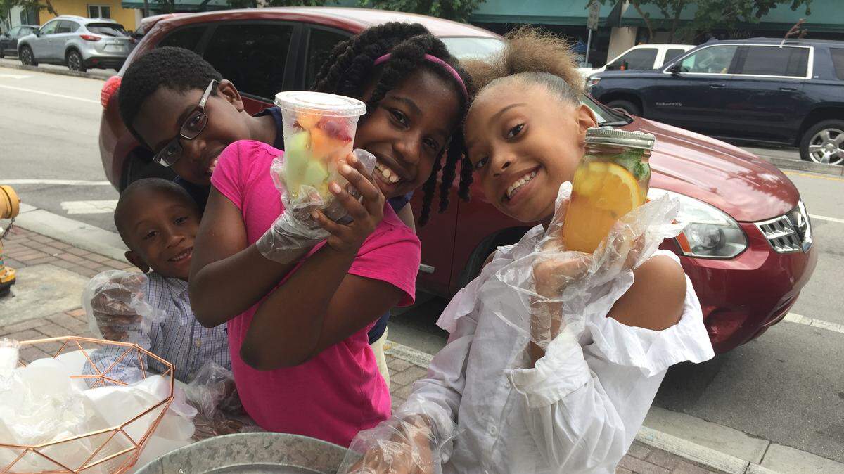 Le’Niyah Heard, in white shirt, Charity Britton, in pink shirt, with Joshua Britton and Nathan Oliver, boy being hugged, are selling cups of fresh fruit and jars of a fruit drink at a pop-up on Grand Avenue in  West Coconut Grove this summer.