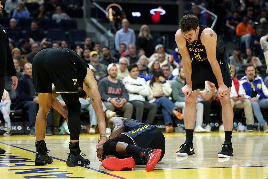 Jimmy Butler III #10 of the Golden State Warriors is looked at by Stephen Curry #30 and Quinten Post #21 after he collided with Davion Mitchell #45 of the Miami Heat and injured his knee during the third quarter at Chase Center on January 19, 2026 in San Francisco.