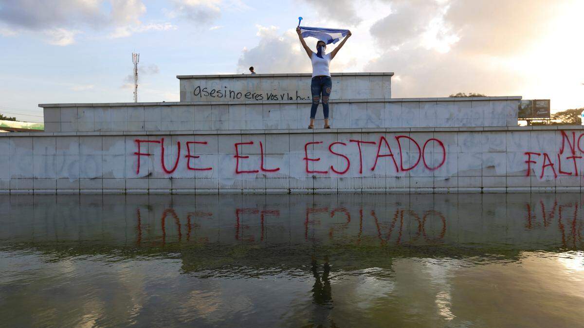 A masked protester holds up a national flag as she stands above graffiti reading in Spanish 'The state did it,' during a protest against the government of President Daniel Ortega on National Children's Day, in Managua, Nicaragua, Friday, June 1, 2018.