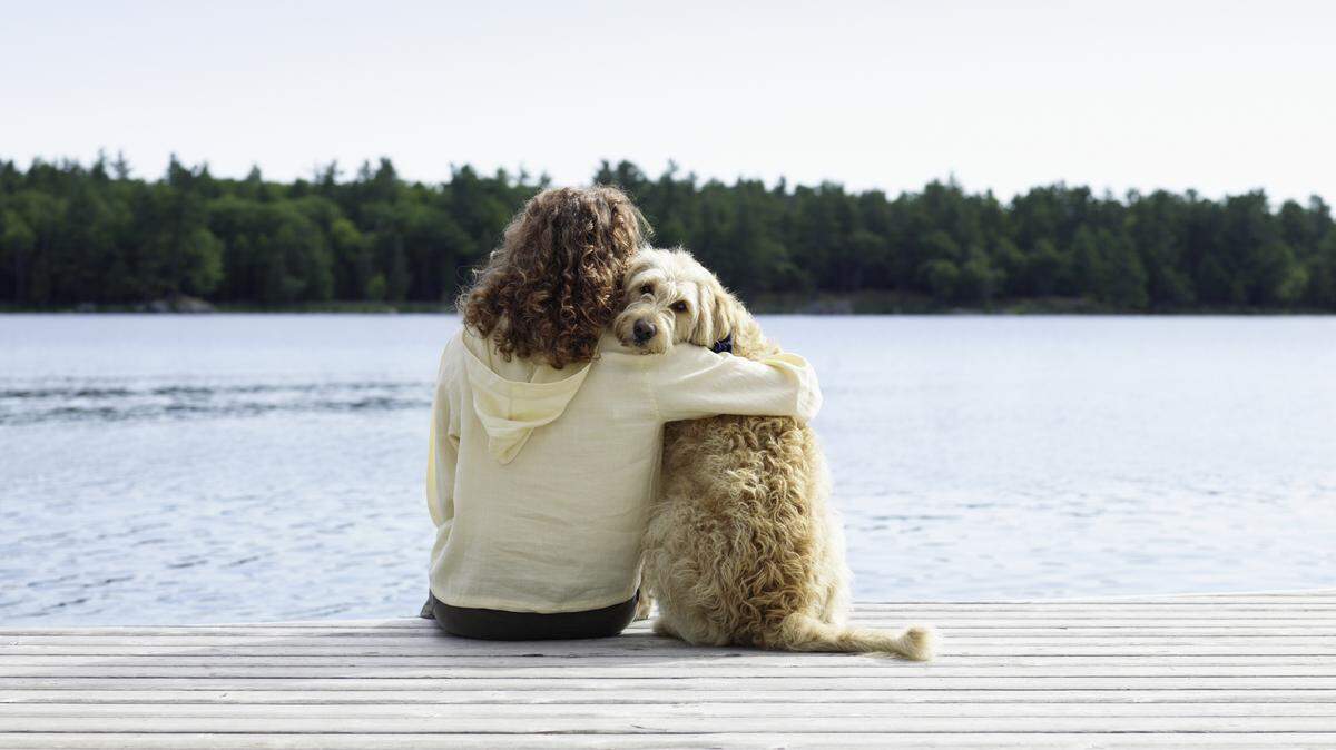 A woman sits on a dock with her arm around her dog. 