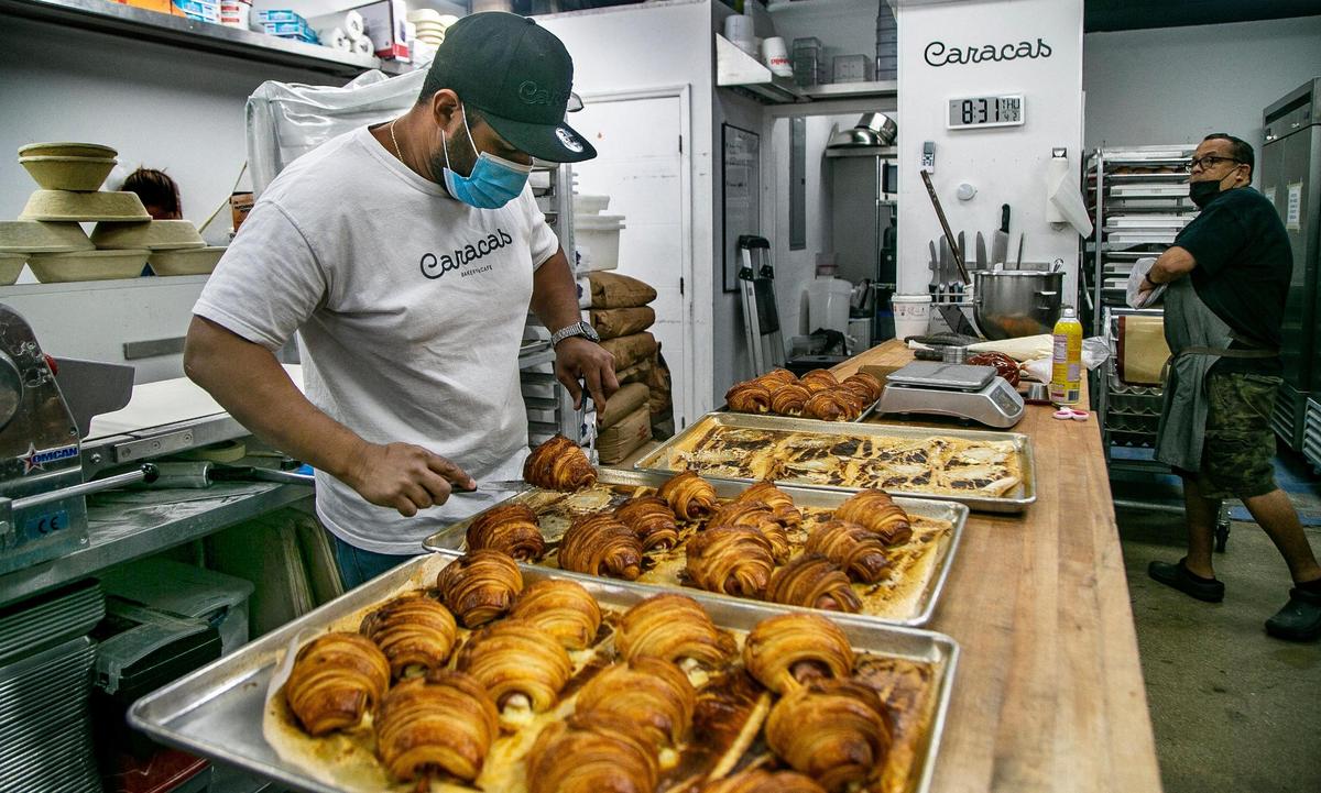 Luis Mauricio Payares checks trays of ham and cheese croissants just out of the oven, at the Caracas Bakery in Doral owned by father and son Manuel, right, and Jesus Brazon, who came from Venezuela years apart, settled in Miami, and opened up the bakery together a year ago, just before the pandemic.